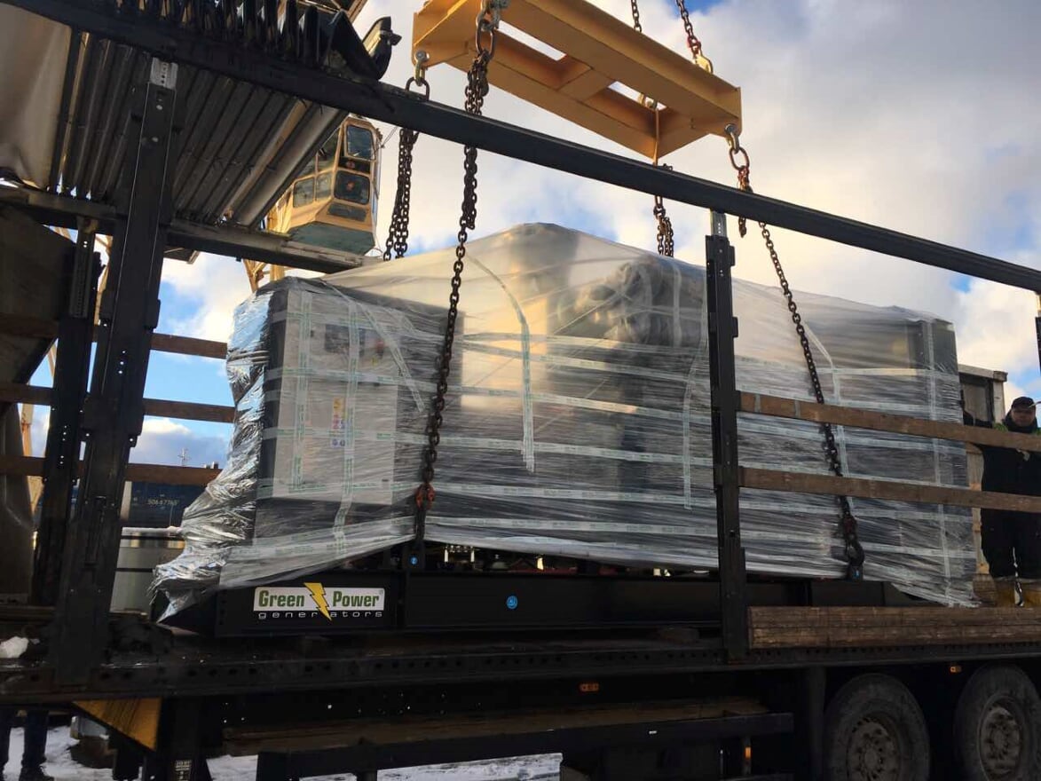 A packaged generator being loaded by crane into an open curtainsider trailer